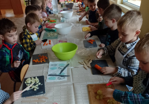 Dzieci siedzą przy stol, kroją warzywa.