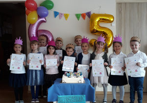Grupa dzieci na tle balonów oraz cyfry 5 i 6, trzymają dyplomy urodzinowe.