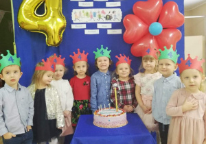 Dzieci obchodzące czwarte urodziny stoja na tle dekoracji przed urodzinowym tortem.